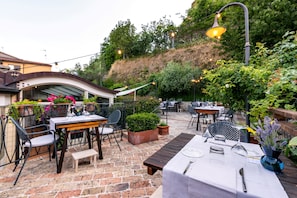 Un espace extérieur aménagé en terrasse, avec des tables et des chaises, entouré de verdure et d’un mur en pierre.