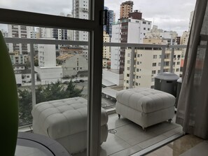 Un balcón con dos sillas blancas acolchadas, vistas a un paisaje urbano con diversos edificios y un equipo de aire acondicionado grande.