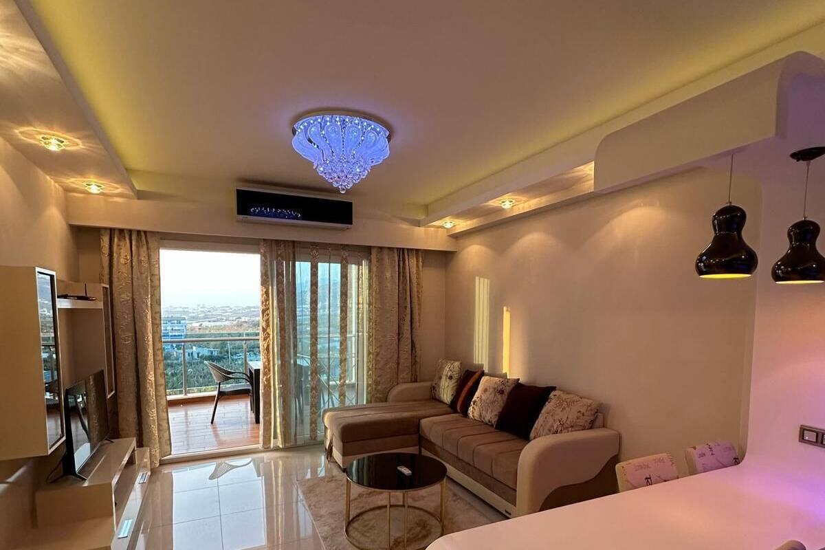 Una sala de estar moderna con sofá, mesa de centro y televisor, ventanas amplias con vista a la ciudad y un candelabro.