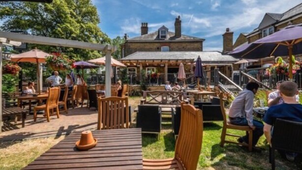 Eine Außenpatio-Terrasse mit Holztischen und -stühlen, Sonnenschirmen und einem Steinhaus im Hintergrund.