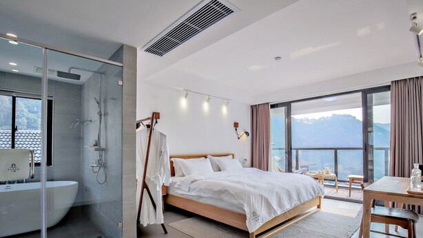 A modern hotel room with a glass-enclosed shower, a large bed with white linens, a wooden desk, and a view of mountains through large windows. - Hangzhou Sansu Hotel (Hangzhou)