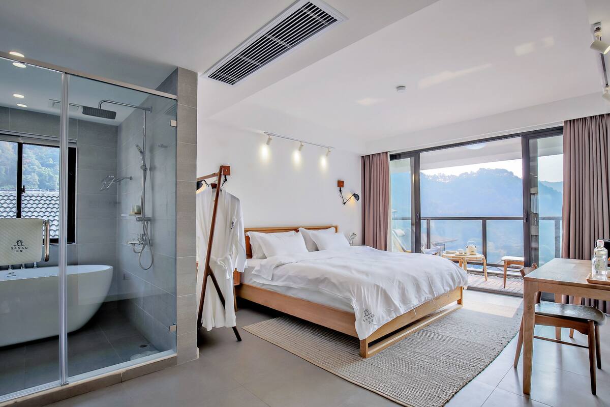 Une chambre d’hôtel moderne dotée d’une douche en verre, d’un grand lit avec du linge de lit blanc, d’un bureau en bois et offrant une vue sur les montagnes grâce à de grandes fenêtres.