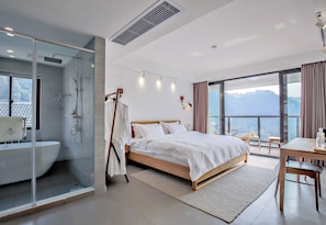 Una habitación de hotel moderna con ducha de vidrio, una cama grande con ropa blanca, un escritorio de madera y vistas a las montañas a través de amplios ventanales.