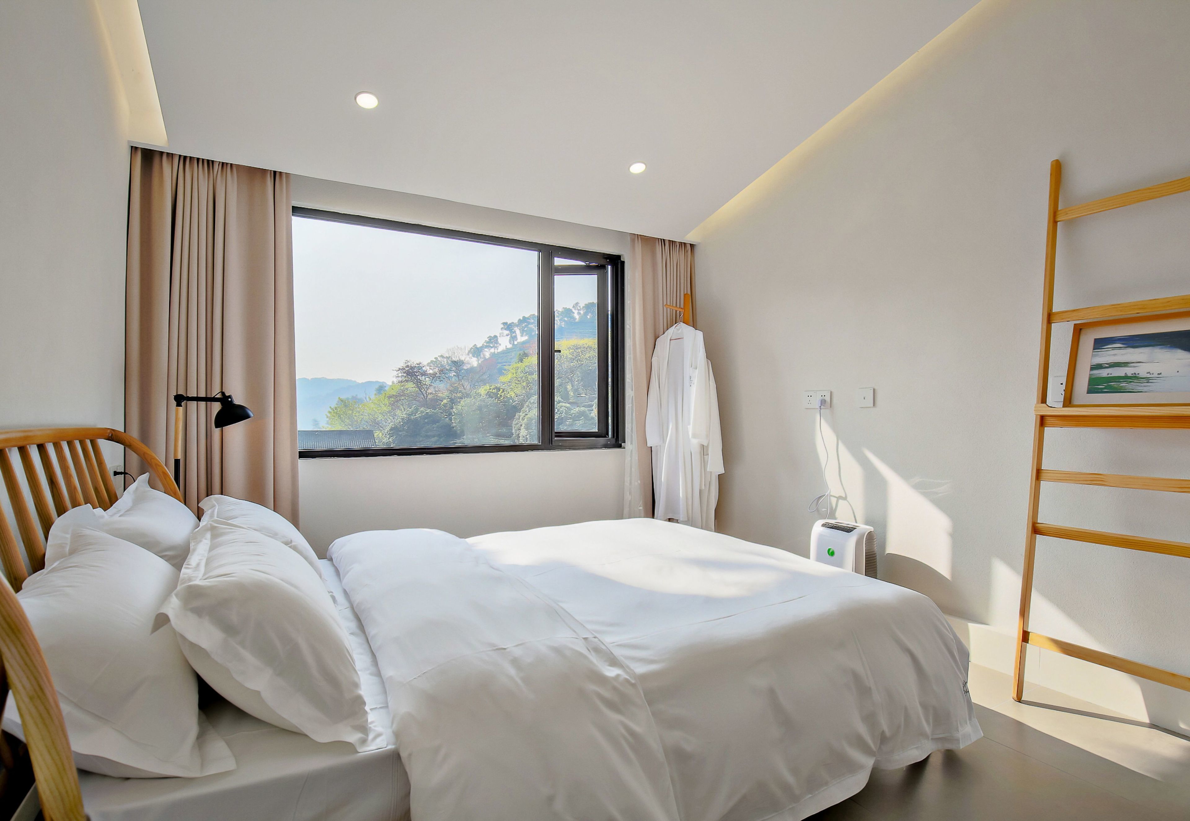 A bedroom with a large window, a bed with white bedding, a wooden chair, a bedside table, and a wall-mounted ladder.