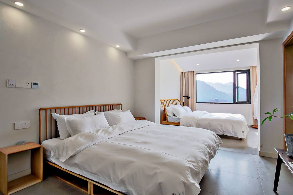 Une chambre d’hôtel moderne avec deux lits, une table de chevet en bois, une petite table et une vue sur les montagnes par la fenêtre.