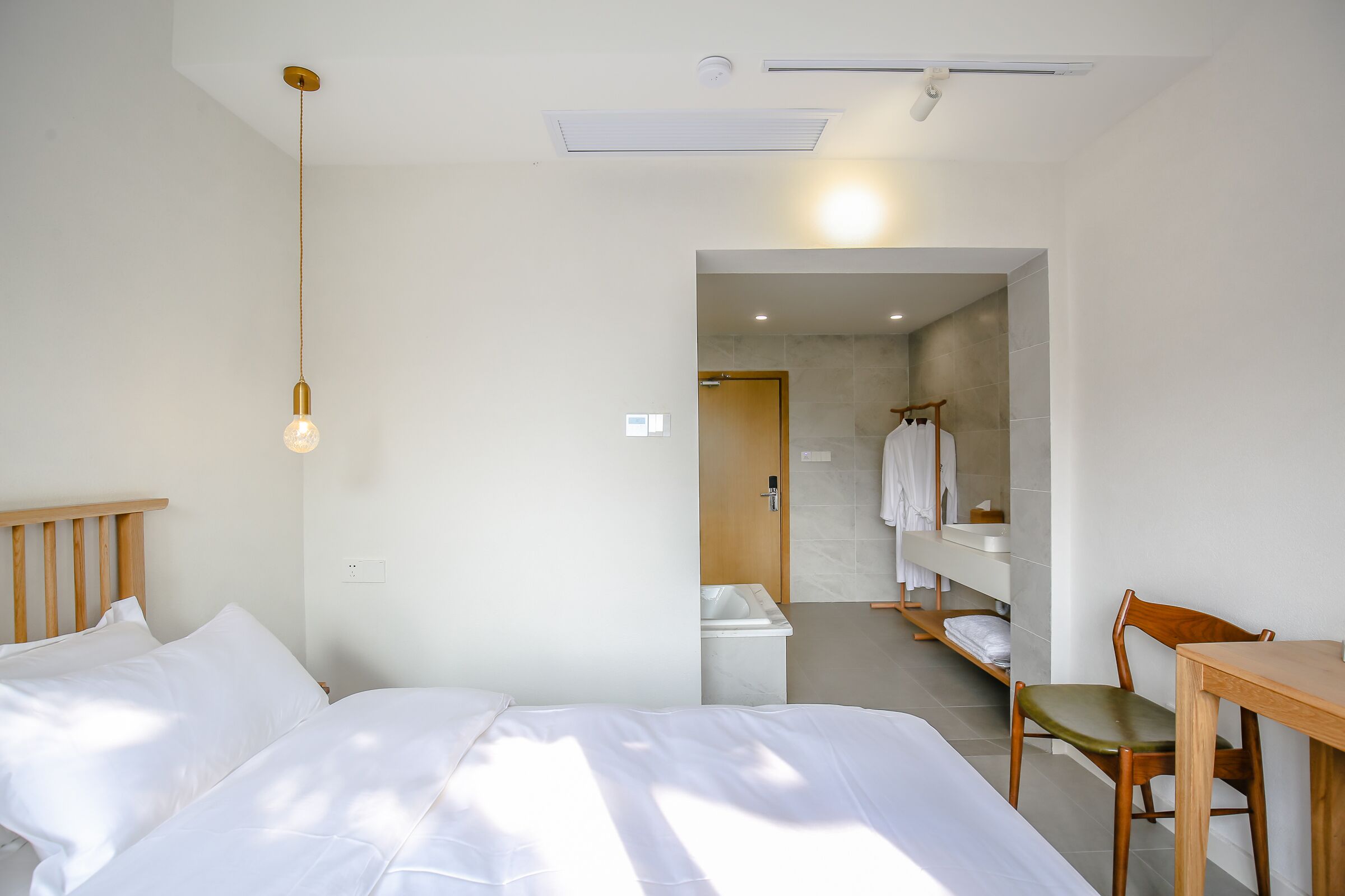Une chambre d’hôtel avec un lit, une chaise en bois, une petite table, un luminaire suspendu et une salle de bain visible à travers une porte ouverte.