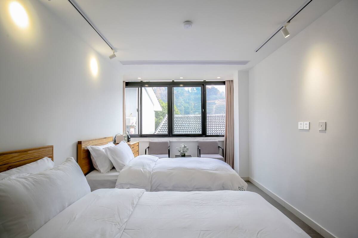 Une chambre d’hôtel avec deux lits, une fenêtre donnant sur des immeubles et une petite table avec un vase.