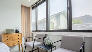 A hotel room with a bed, a wooden dresser, two armchairs, a small table with a plant, and a large window with a view of buildings and trees. - Hangzhou Sansu Hotel (Hangzhou)