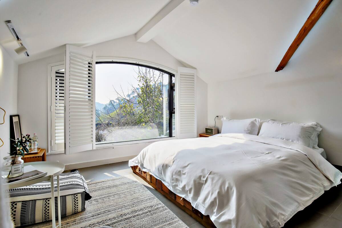 Une chambre avec un grand lit, une fenêtre avec des volets, une table de chevet et une vue sur les arbres à l’extérieur.