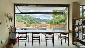 A modern interior with a large window overlooking a mountainous landscape. Inside, there is a dining area with chairs and a table, and a bookshelf filled with books. - Hangzhou Sansu Hotel (Hangzhou)
