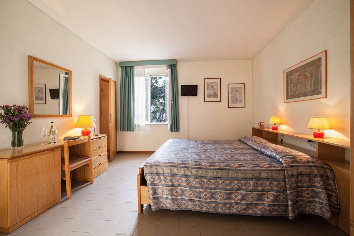 A bedroom with a bed, a desk with a lamp, a dresser, a mirror, and a window with curtains.