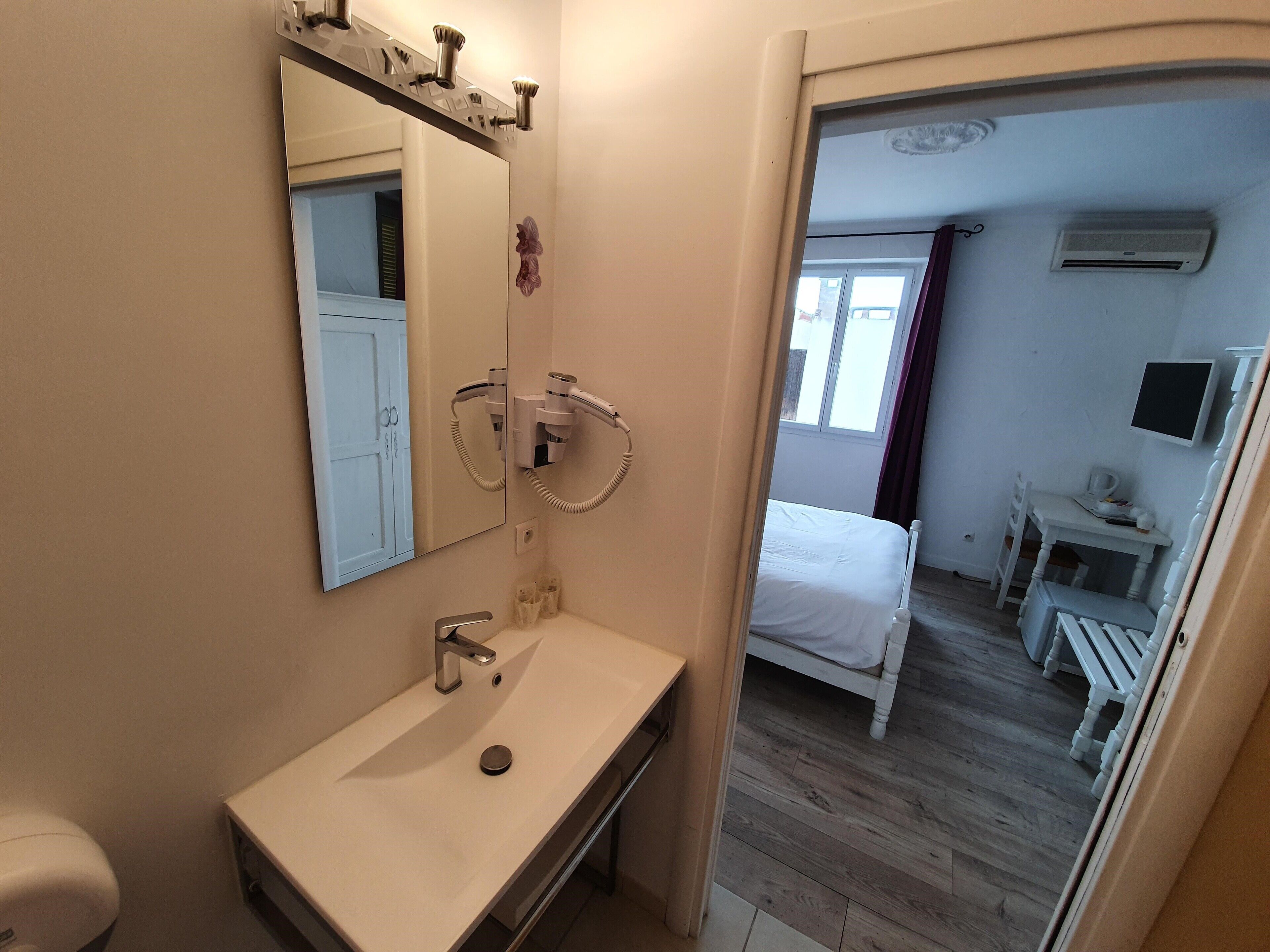 A bathroom with a sink, mirror, and hairdryer. There is a bed and a desk with a chair in the adjacent room.