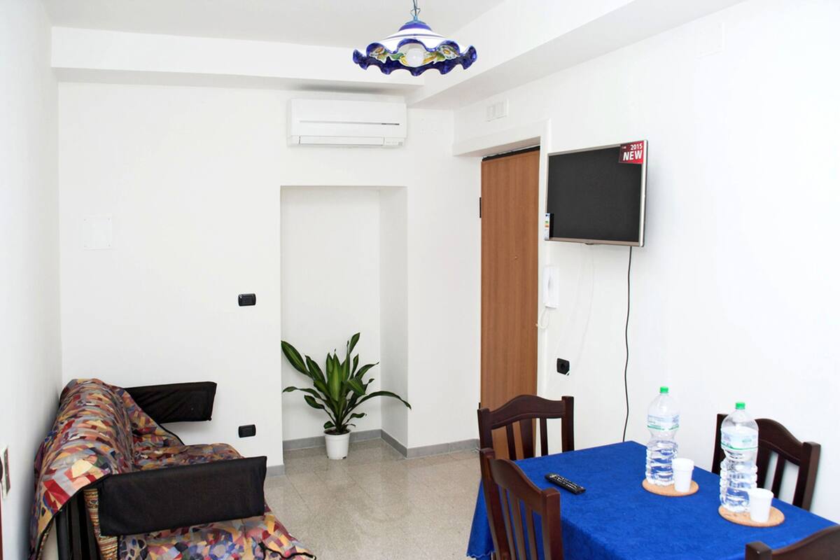 A room with a bed, a dining table with chairs, a potted plant, a television, and a water dispenser.