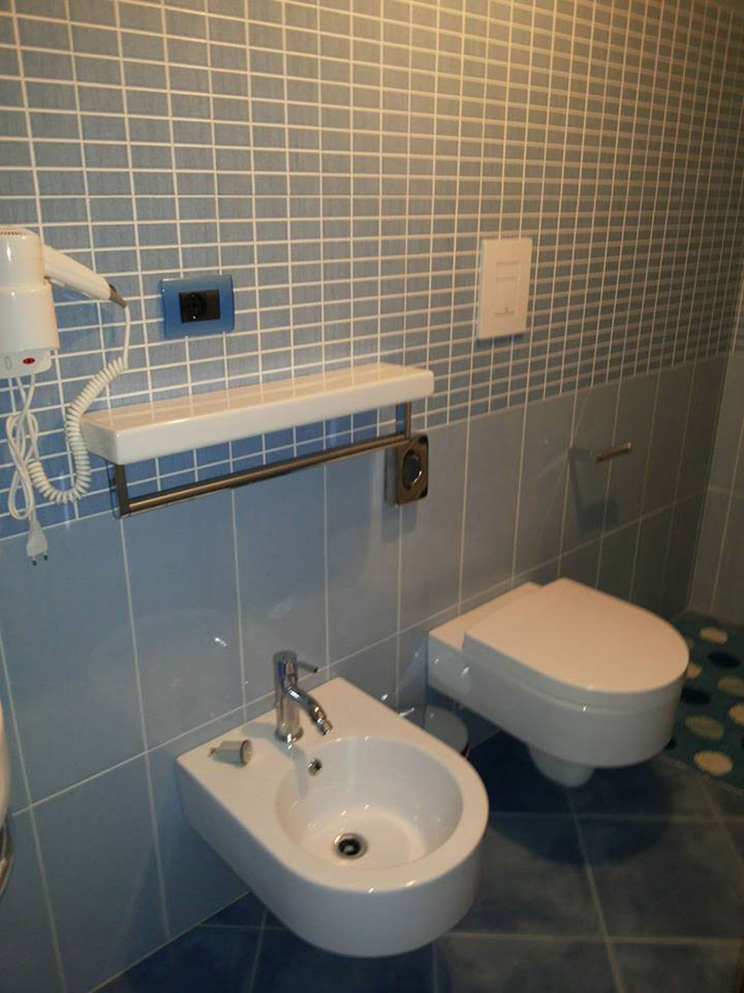 A bathroom with a bidet, a toilet, a towel rack, and a hairdryer on the wall.