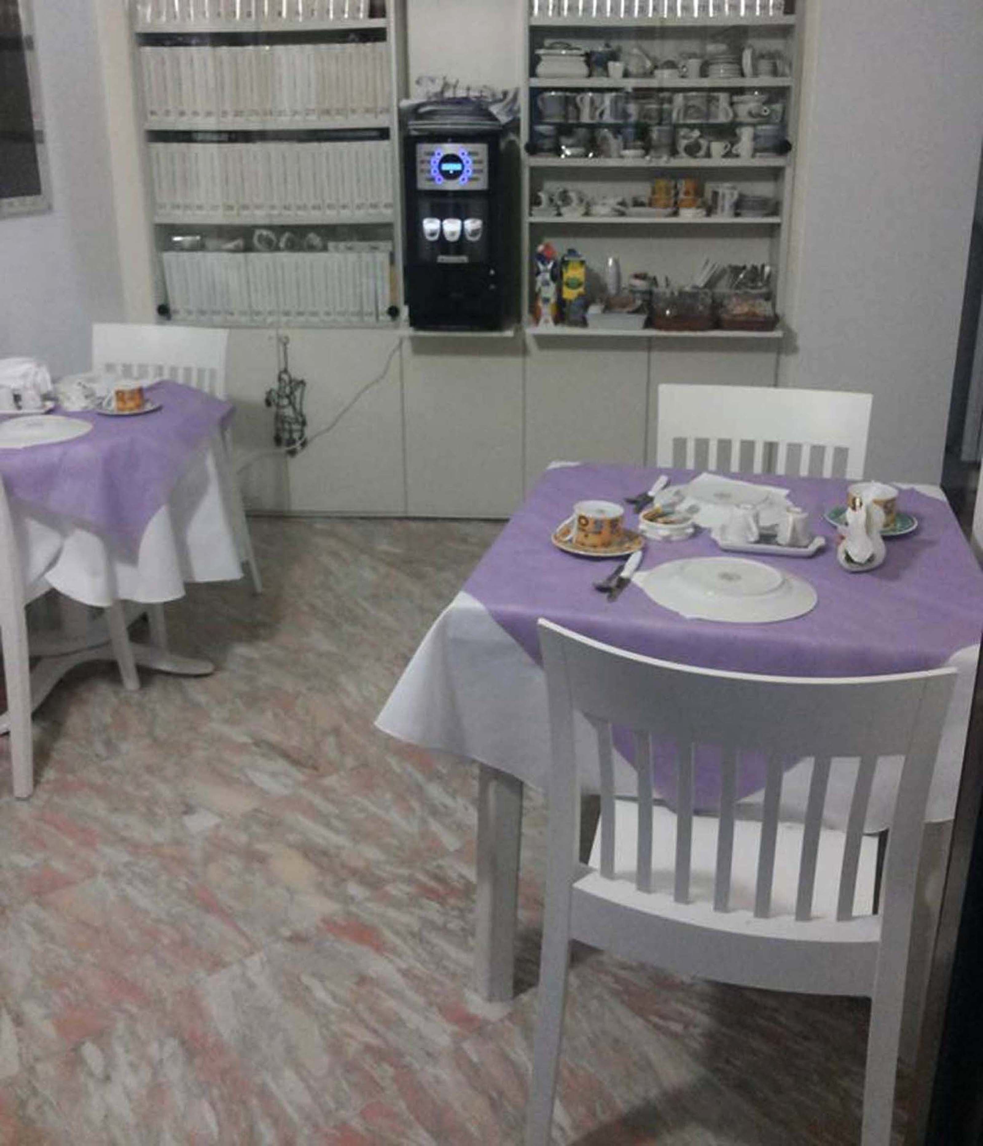 Un coin repas avec deux tables, chacune recouverte d’une nappe violette. Des chaises sont disposées autour des tables, et une machine à café est visible au fond.