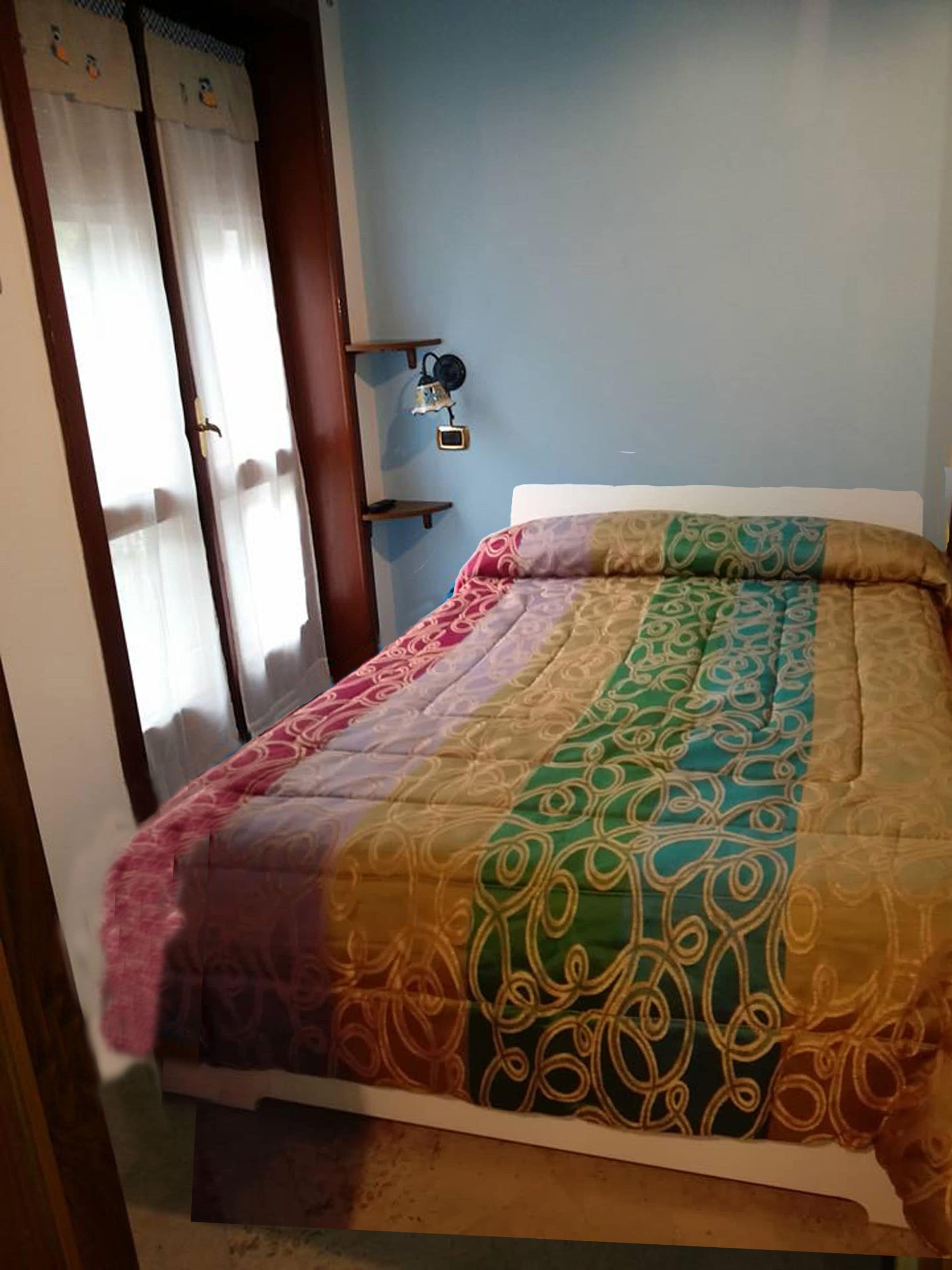 A bed with a multicolored quilt, a wooden door with a curtain, and a wall-mounted light fixture.