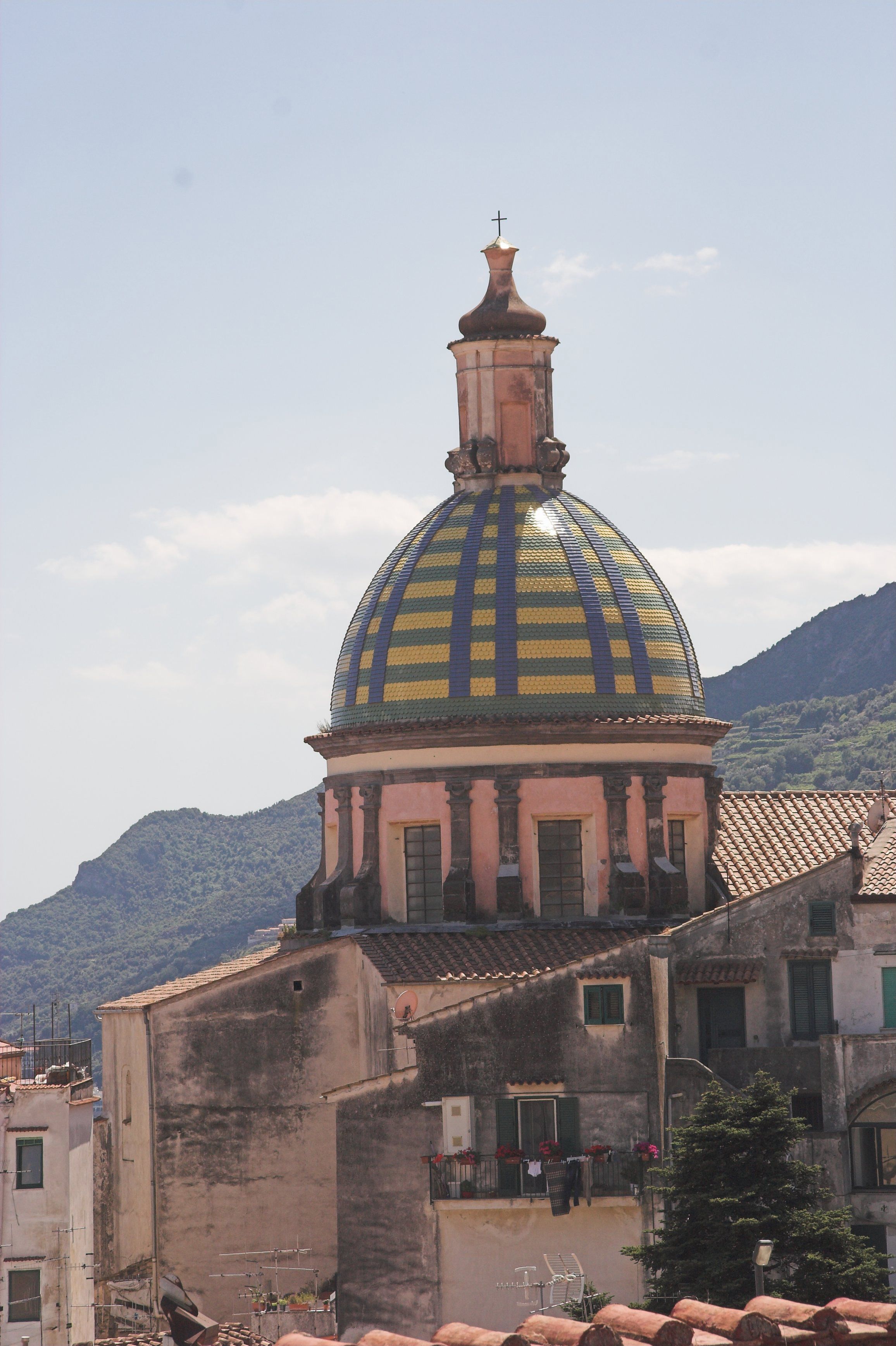 Un edificio con una cúpula destacada y una cruz en la parte superior, rodeado de otras construcciones y un paisaje montañoso al fondo.