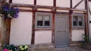 Ein traditionelles Fachwerkhäuschen mit einem pflasterbelagten Eingang, zwei Fenstern mit Fensterläden und einer Tür.