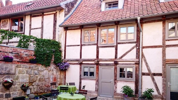 Ein traditionelles Fachwerkhäuschen mit Innenhof. Man sieht eine Steinmauer, eine Holztür und einen kleinen Tisch mit Stühlen.