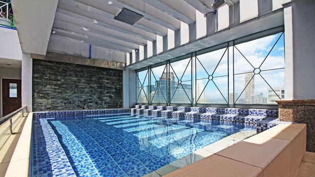 A swimming pool with blue mosaic tiles, a stone wall, and a view of city buildings through large windows. - BSA Mansion (Makati)