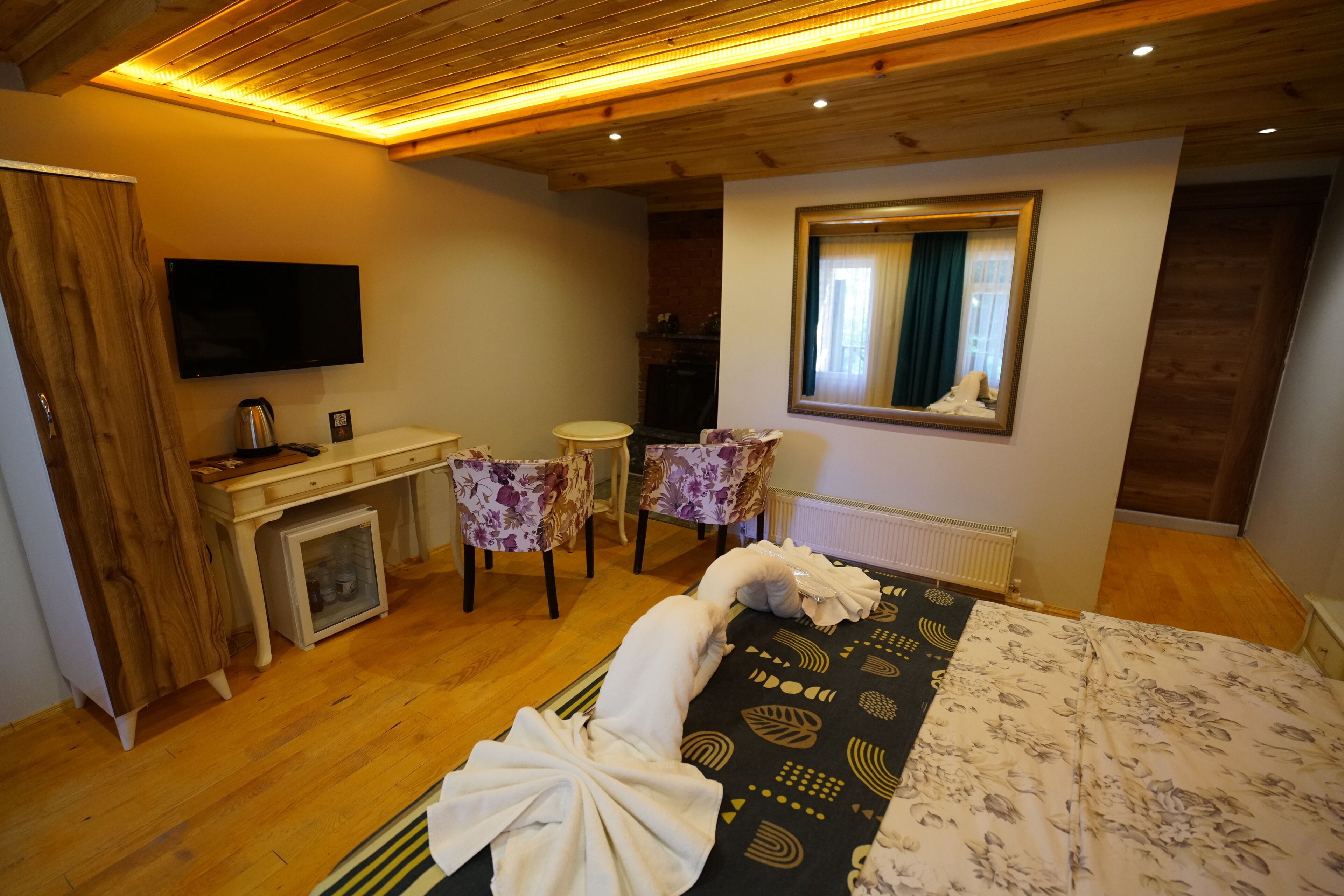Habitación de hotel con techo de madera, cama con colcha estampada, escritorio con televisor de pantalla plana, chimenea y dos sillones tapizados con motivos florales.