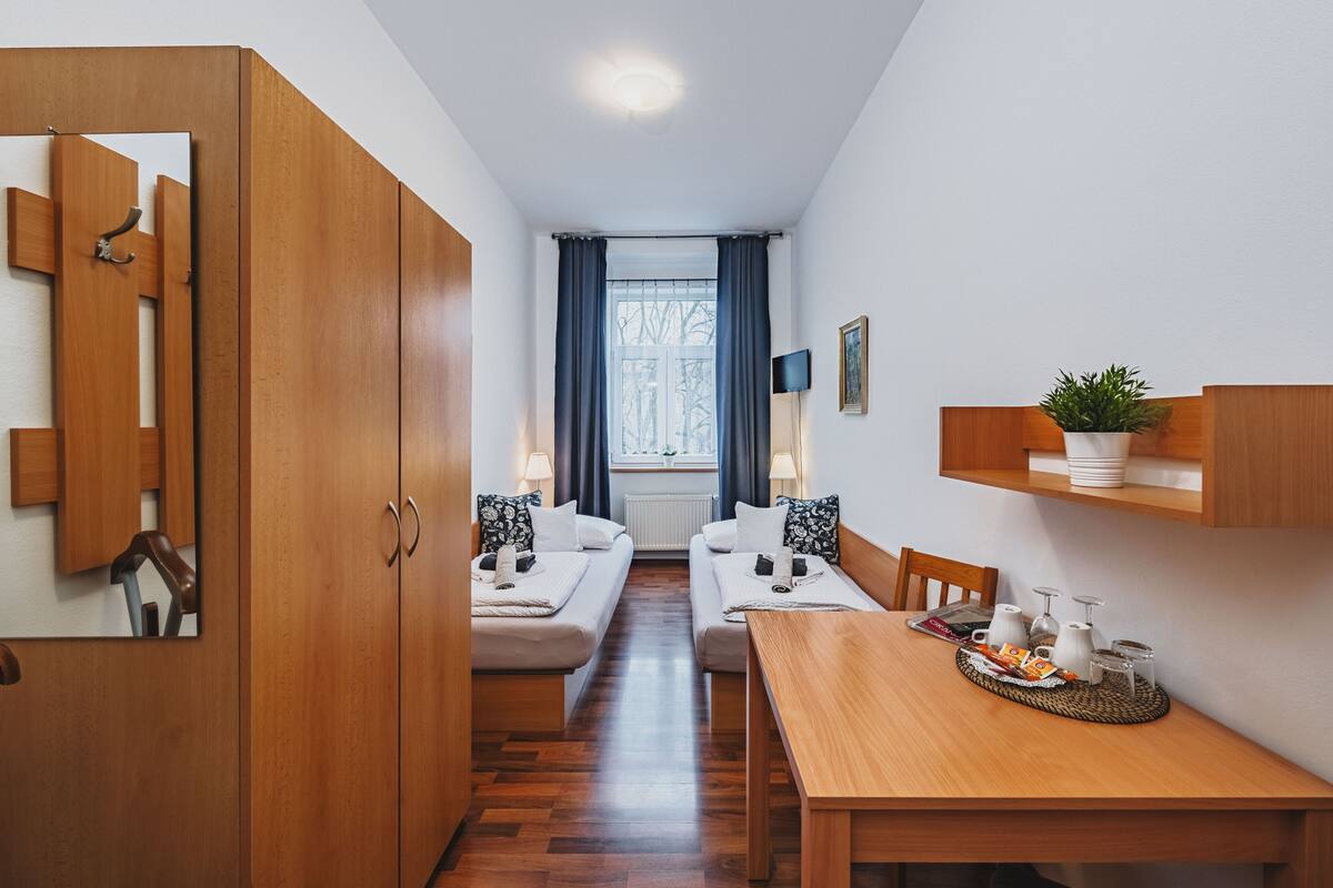 A modern hotel room with a wooden wardrobe, a dining table with a breakfast tray, two sofas, a window with curtains, and a small plant on a shelf.