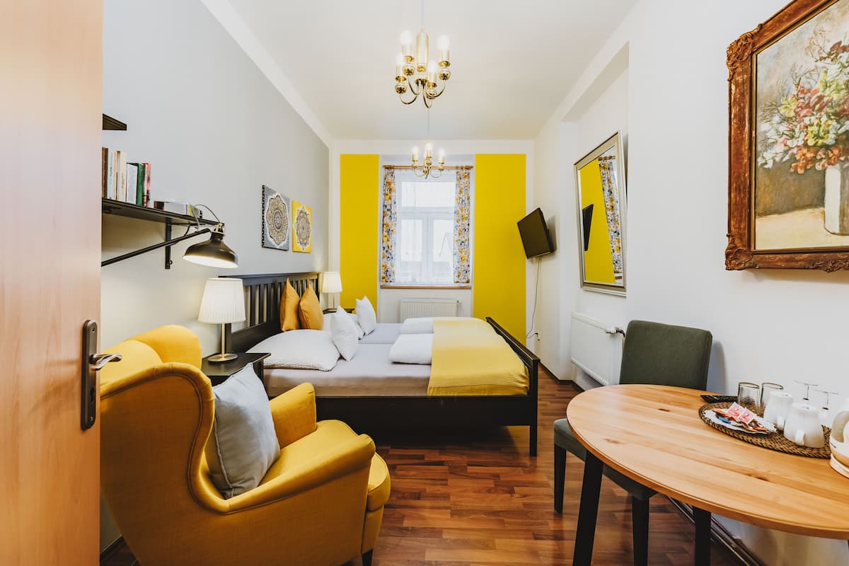 A hotel room with a bed, a yellow armchair, a wooden table with tea cups, a lamp, and a painting on the wall.