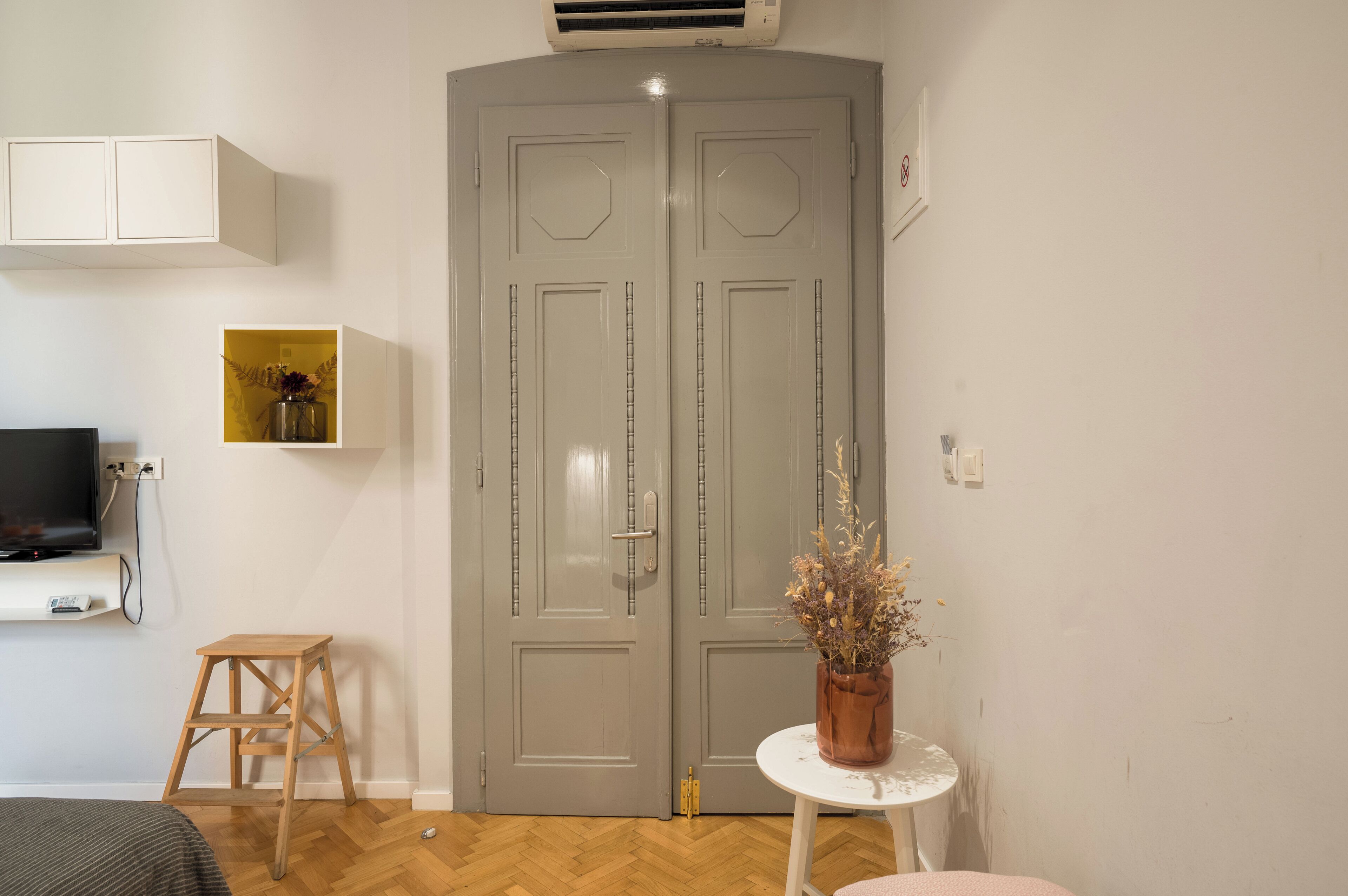 A room with a grey double door, a small wooden stool, a white table with a vase, and a mounted television.