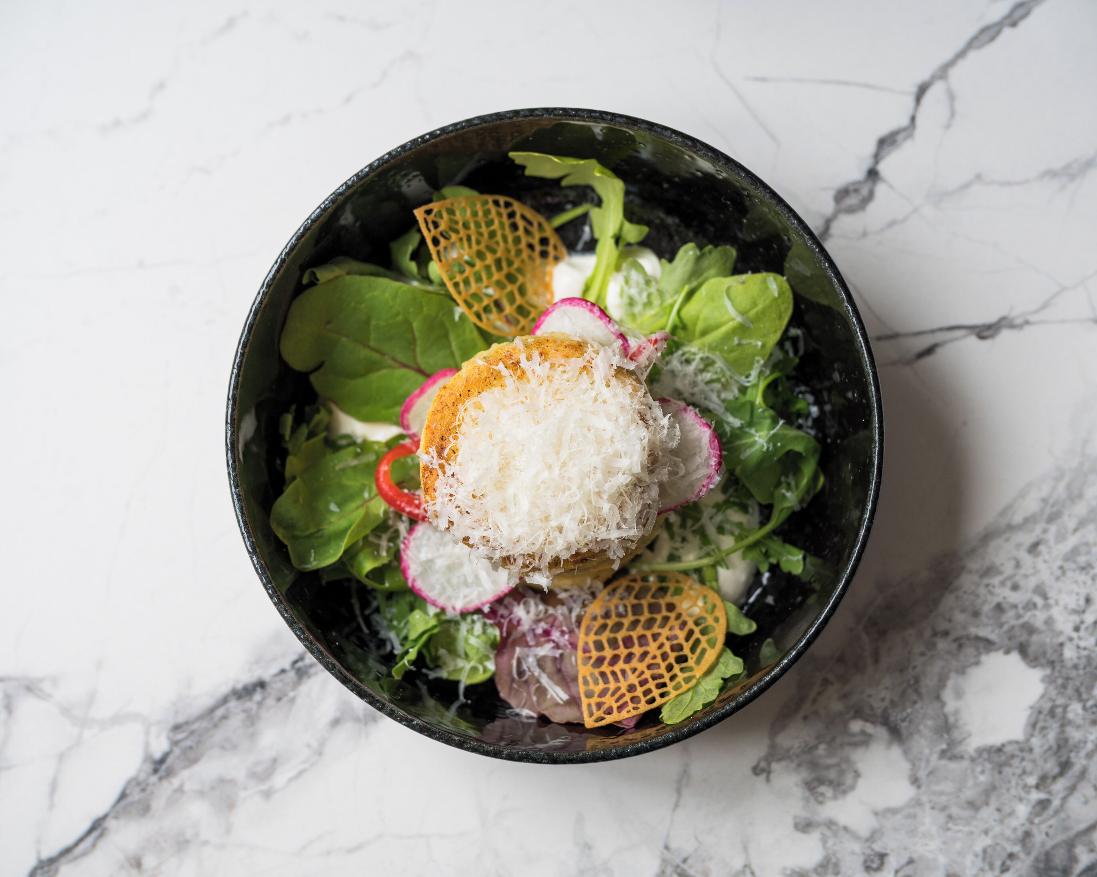 A bowl of salad with various greens, radishes, and shredded cheese on a marble surface.