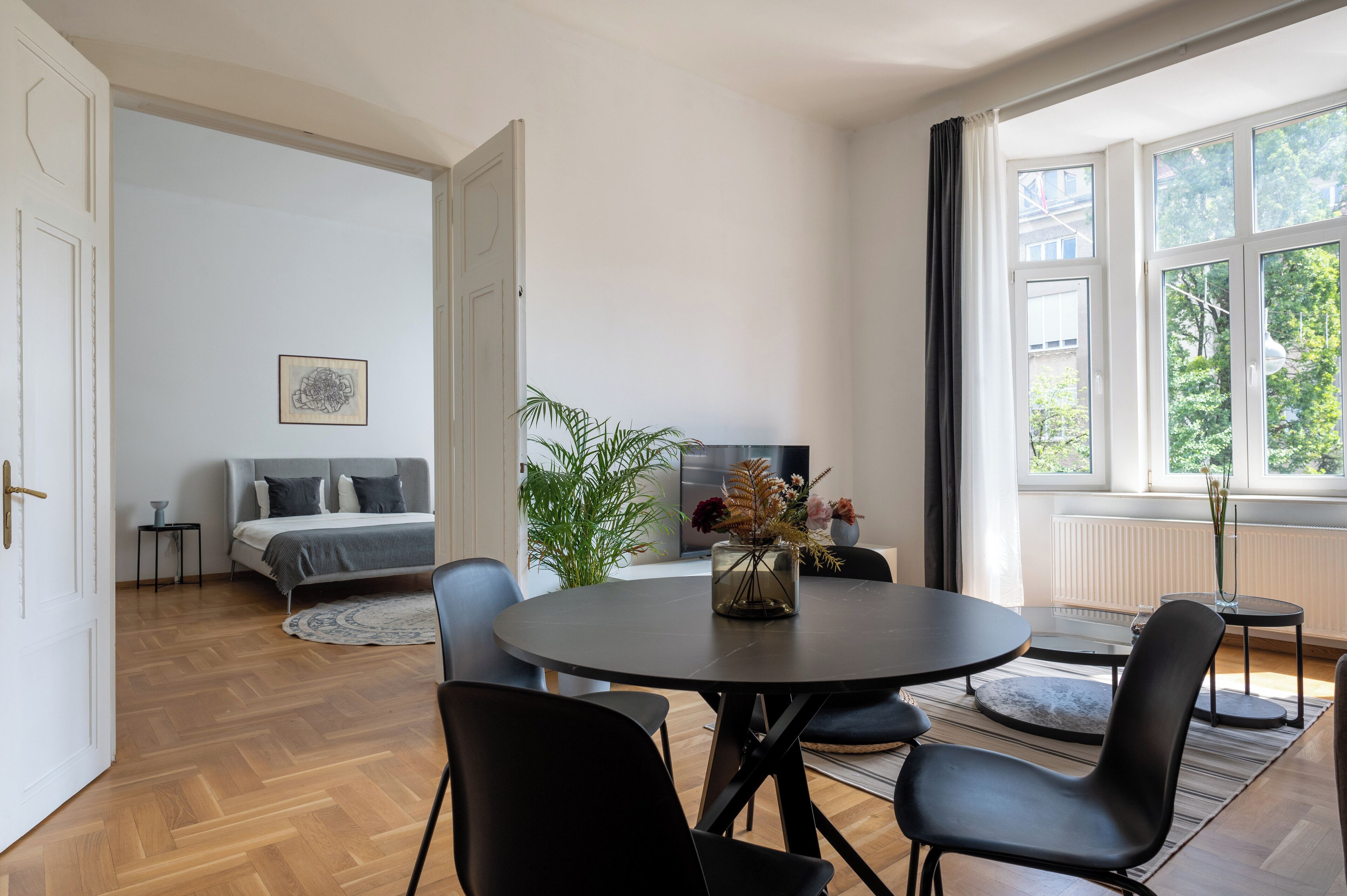 Ein modernes Wohnzimmer mit einem runden Esstisch, schwarzen Stühlen, einem grauen Sofa und einem großen Fenster mit Vorhängen.