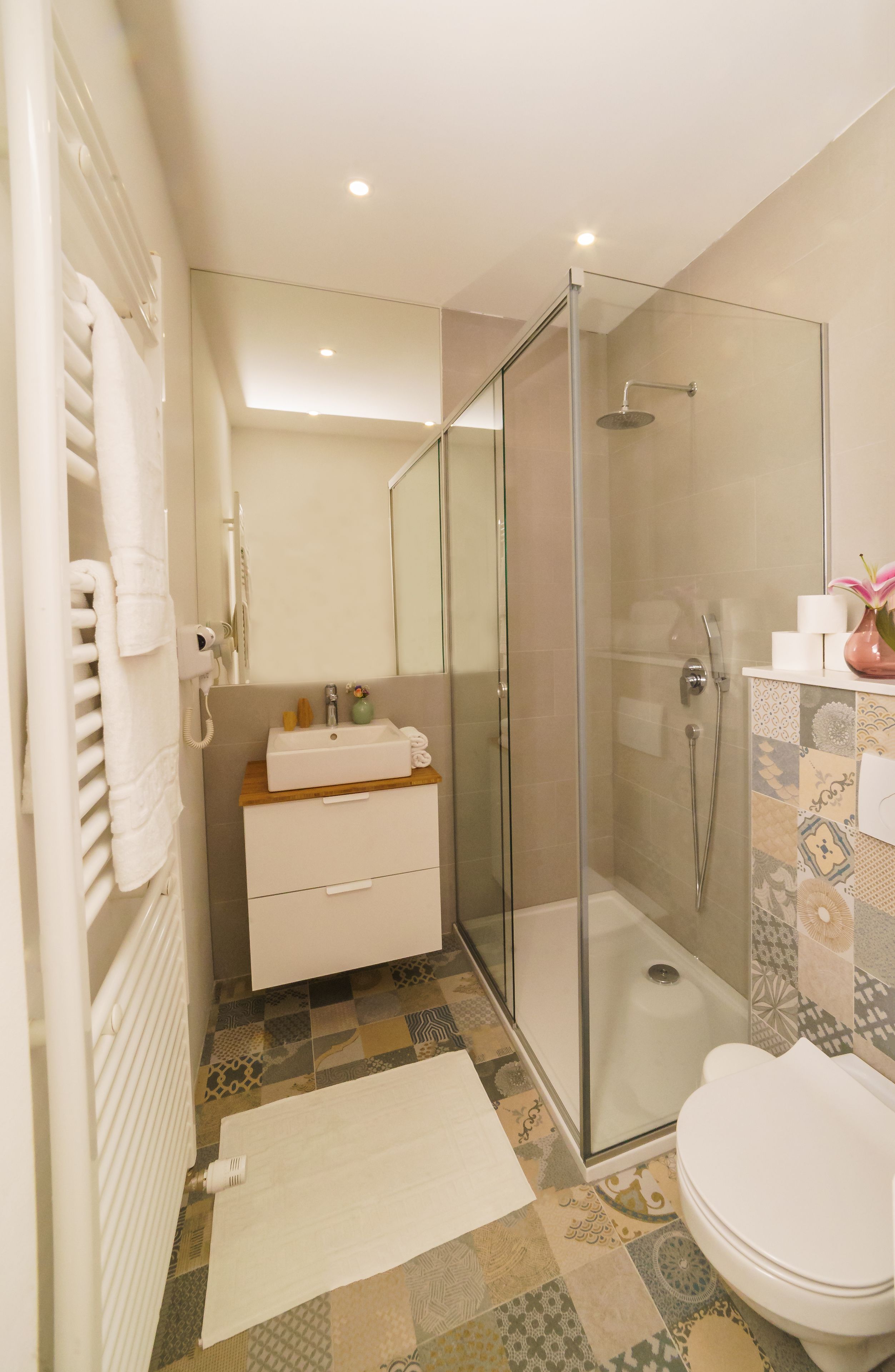 Un baño moderno con ducha envuelta en vidrio, un lavabo blanco con encimera de madera y suelo estampado.