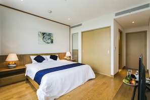 Une chambre d’hôtel avec un grand lit, une tête de lit en bois, deux lampes de chevet, un tableau au mur, une armoire et un plateau avec des fruits et une théière posé sur le sol.