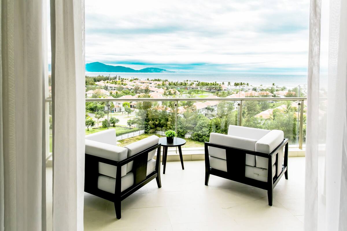 Ein Balkon mit zwei weißen Sofas und einem kleinen Tisch mit Blick auf eine Küstenstadt und Berge.