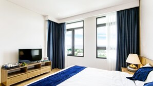 A modern hotel room with a large bed, a flat-screen TV on a wooden console, a bedside table with a lamp, and large windows offering a view. - OCEAN Apartment 2 Bedroom -SABINA DaNang (Da Nang)