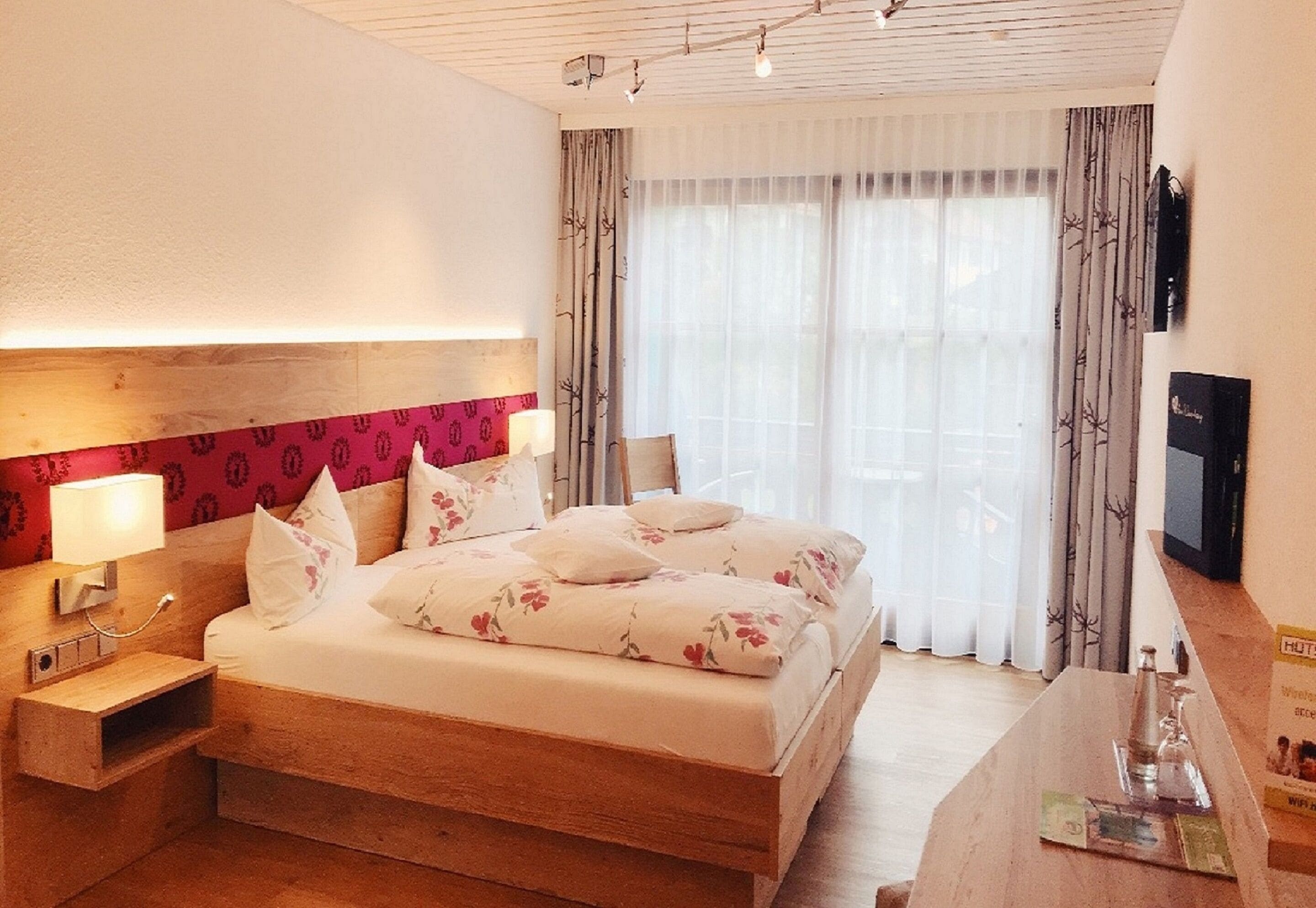 Une chambre d’hôtel avec un lit en bois, une table de chevet avec une lampe, une télévision et une grande fenêtre avec des rideaux.