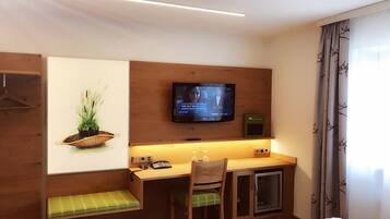A compact room with a wooden desk, a television mounted on the wall, a small bench, and a cabinet.