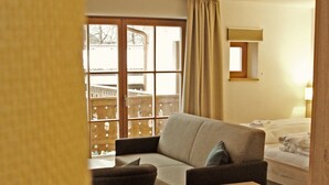 A cozy living room with a sofa, a small wooden side table, and a chair. There is a window with curtains, a wooden floor, and a bed in the background. - Mammhofer Suite & Breakfast (Oberammergau)