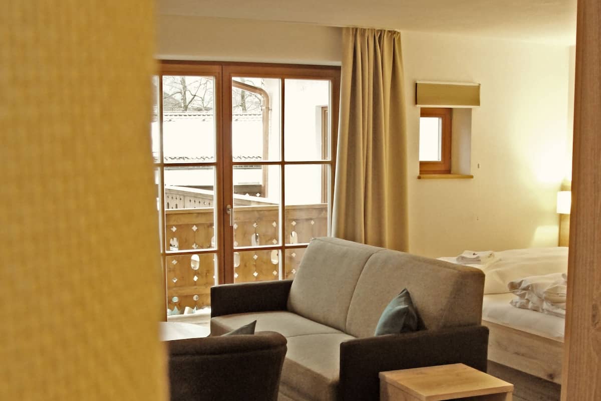 A cozy living room with a sofa, a small wooden side table, and a chair. There is a window with curtains, a wooden floor, and a bed in the background.