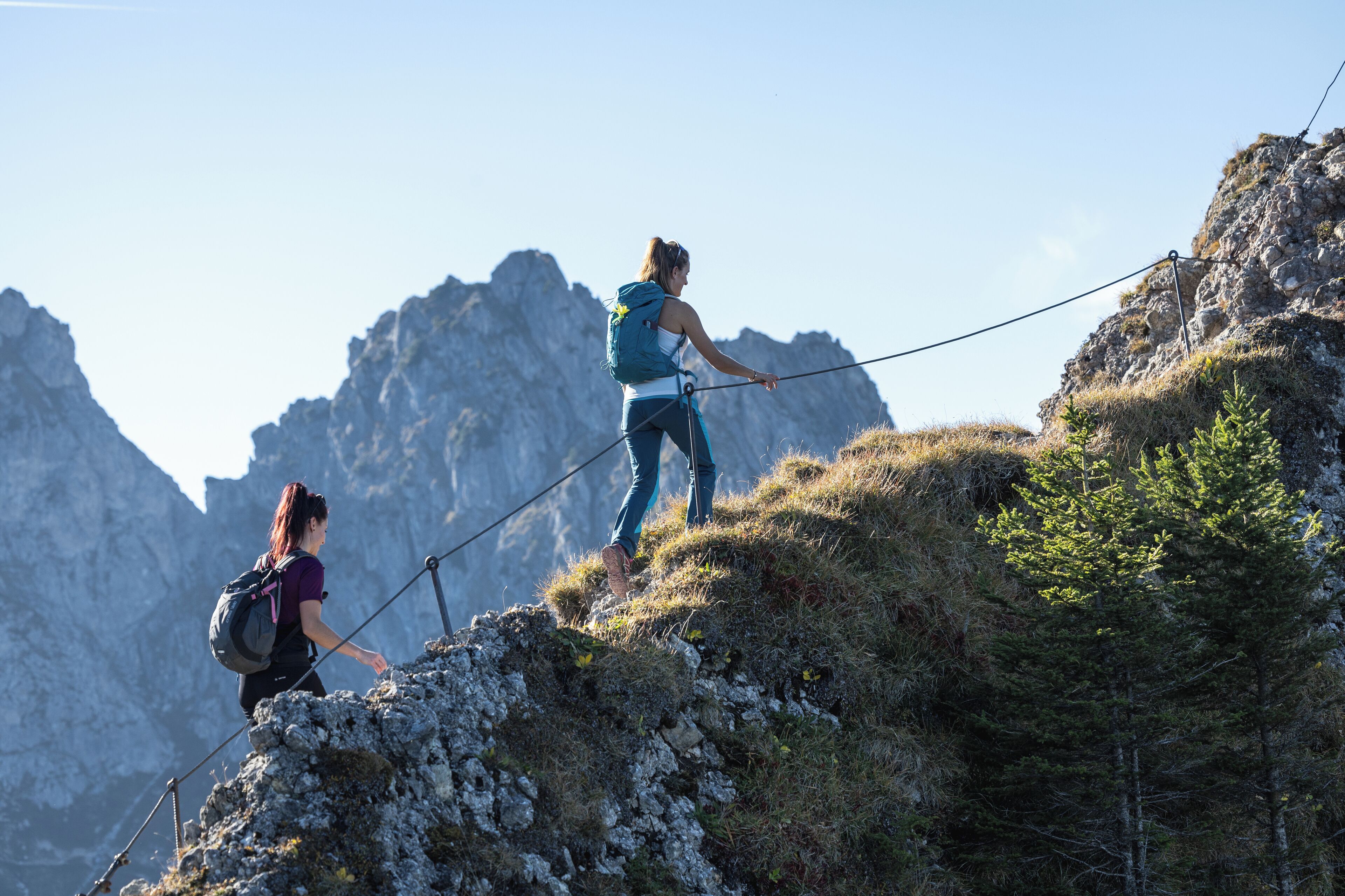 Una persona caminando por un sendero rocoso en la montaña, con cielo despejado y montañas al fondo.