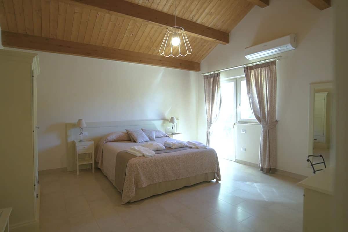 Een slaapkamer met een groot bed, een nachtkastje, een lamp, een plafondlamp en een raam met gordijnen.
