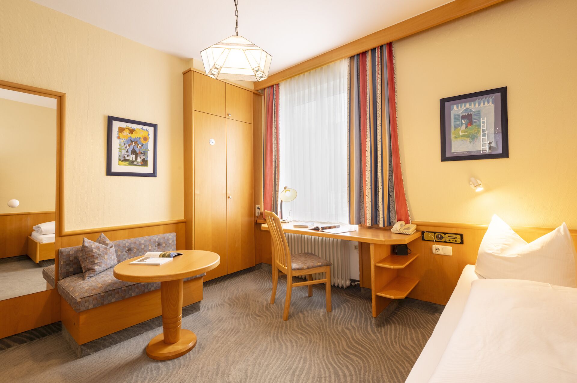A hotel room with a bed, a desk with a chair, a sofa, a radiator, a lamp, and a window with curtains.
