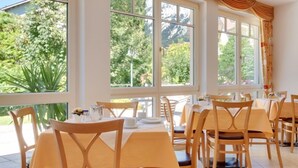 A dining area with multiple tables and chairs, large windows with curtains, and a view of greenery outside.