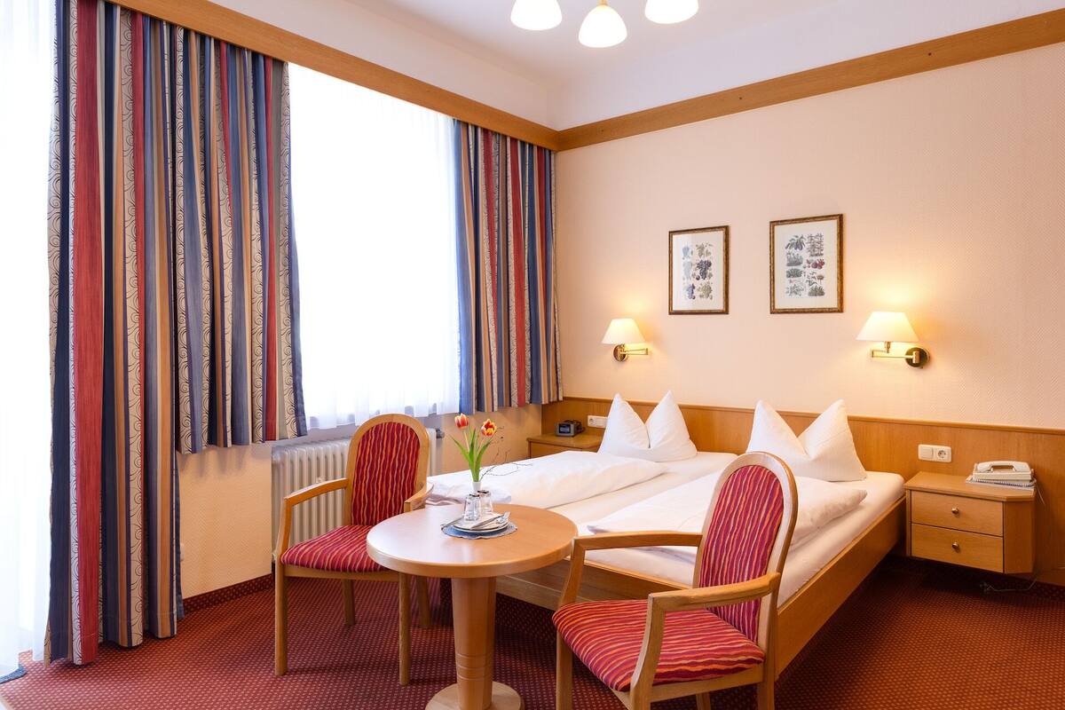 Quarto de hotel com cama, duas cadeiras, uma mesinha, criado-mudo e duas luminárias fixadas na parede.