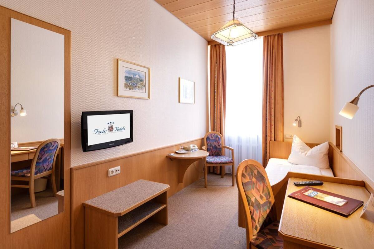 Quarto de hotel com televisão, escrivaninha, cadeira, cama e janela com cortinas.