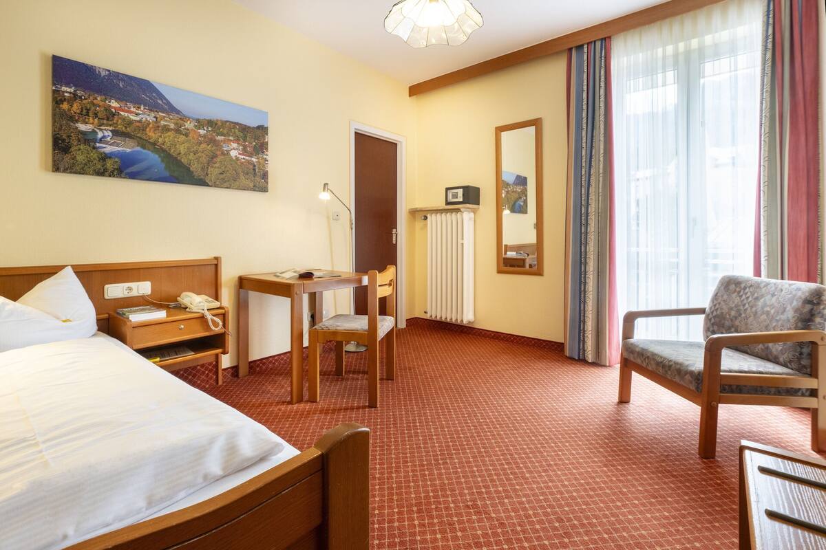 Quarto de hotel com cama, escrivaninha, cadeira, espelho e janela com cortinas.