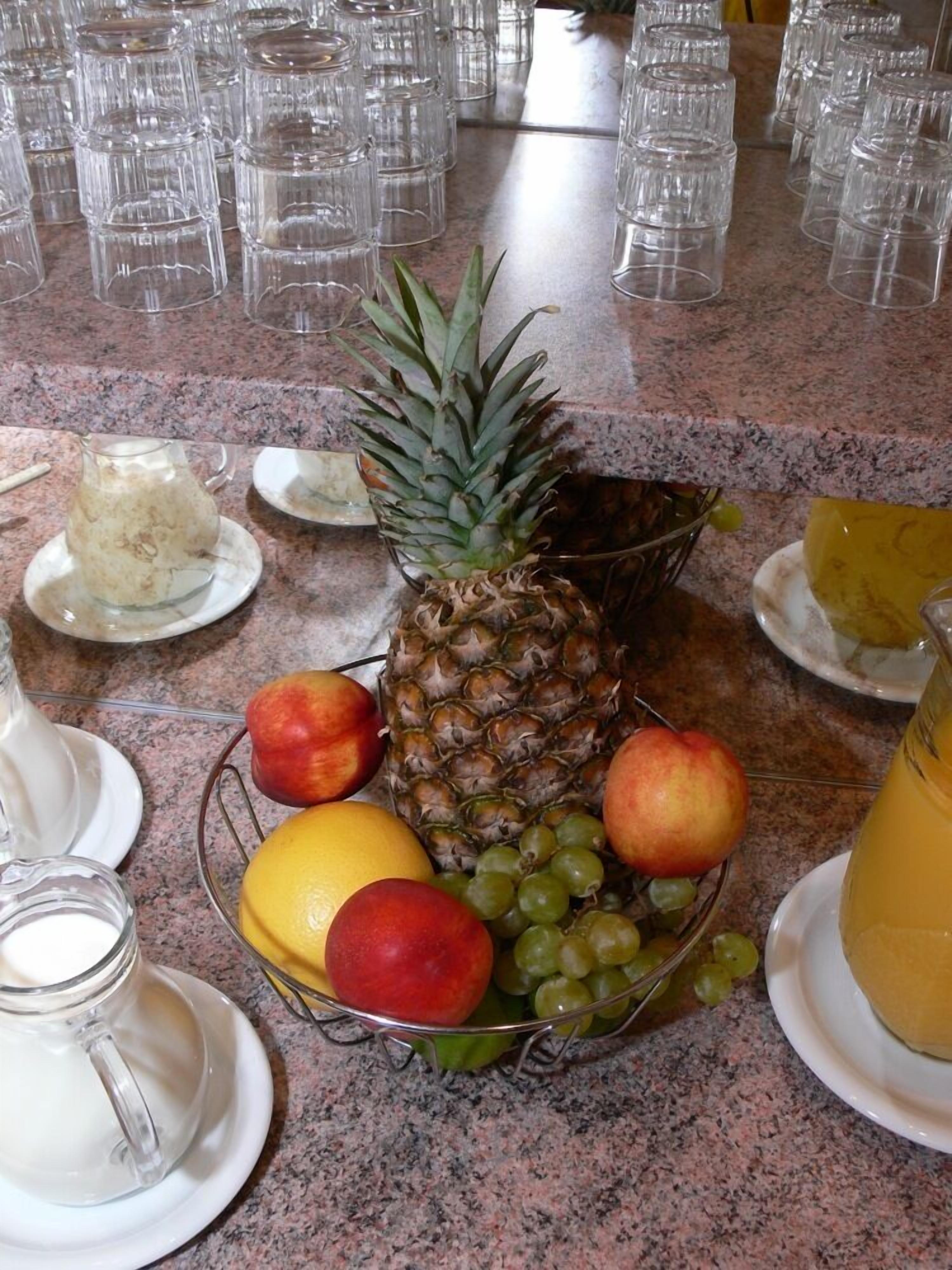 A kitchen counter with a fruit bowl containing a pineapple, apples, grapes, and an orange. There are also several empty glasses and a pitcher of juice.