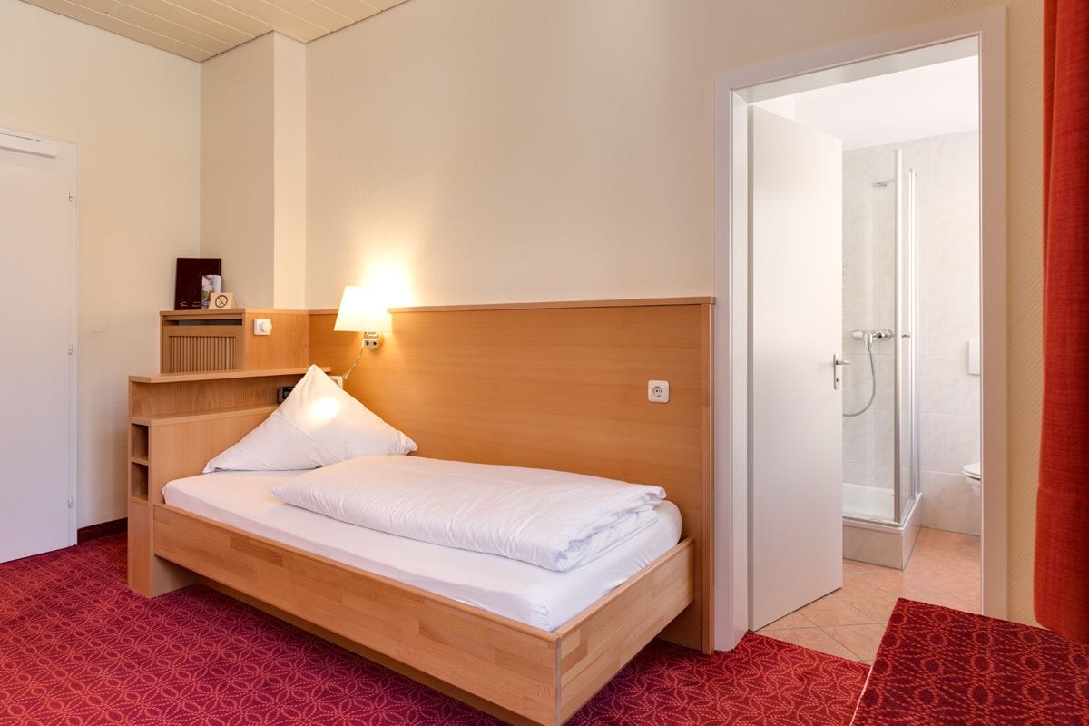 Una cama bien hecha en una habitación de hotel con cabecera de madera, una mesita de noche con lámpara y una alfombra roja estampada.
