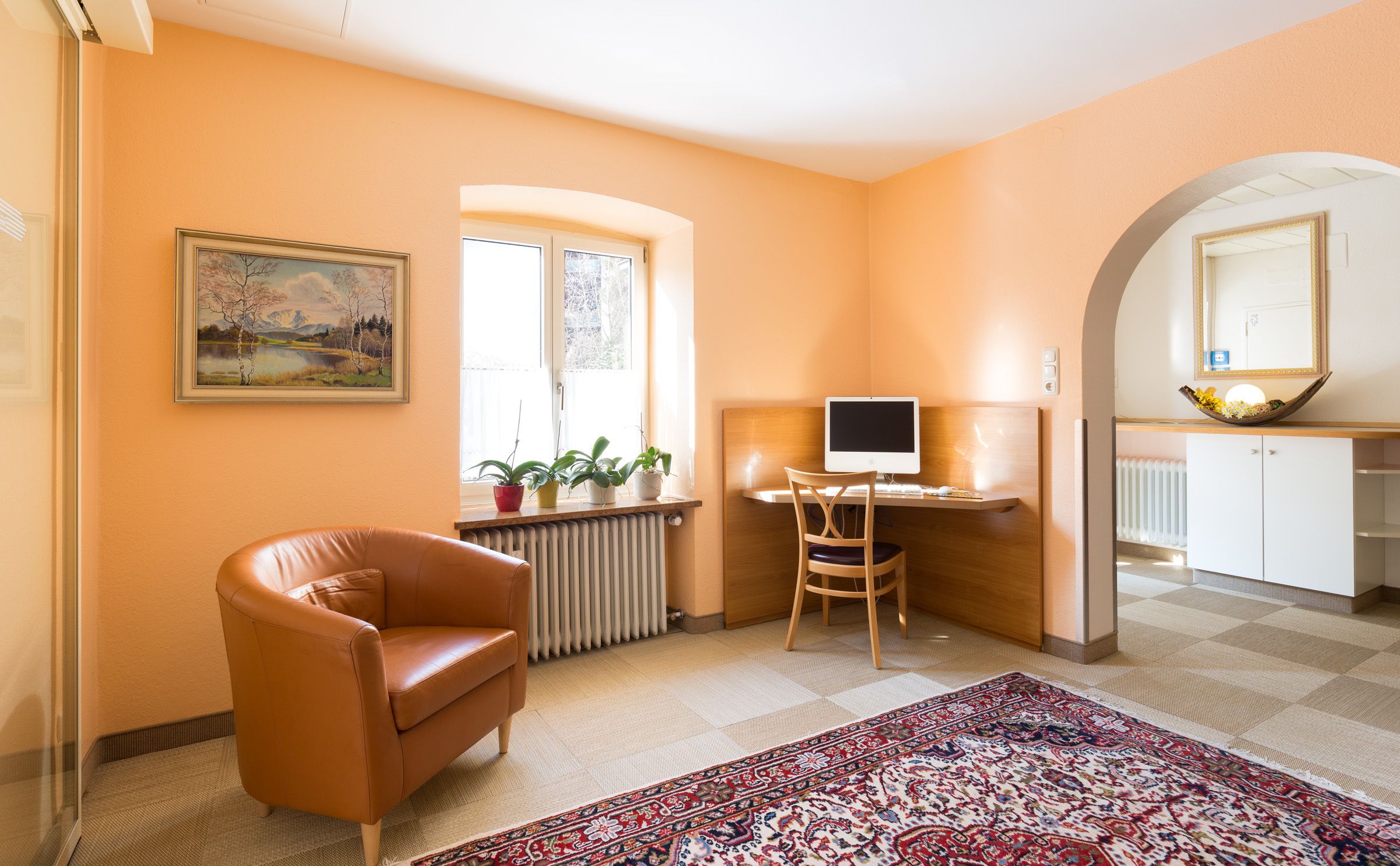 Een gezellige kamer met een leren fauteuil, een bureau met een computer, een radiator en een raam met planten.