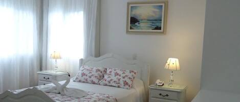 A bedroom with a bed, two bedside tables, a chandelier, a painting, and a window with curtains.