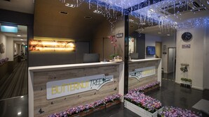 Un mostrador de recepción con detalles de madera y un cartel que dice "BUTTERNUT TREE Hotels". Hay arreglos florales a cada lado del mostrador, y el techo está decorado con luces colgantes.